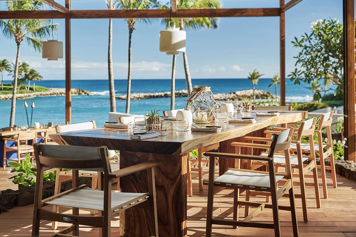 Ein elegant gedeckter, langer Holztisch im Open-Air-Restaurant des Four Seasons Oahu direkt an der Küste mit freiem Blick auf den blauen Pazifik unter Palmen