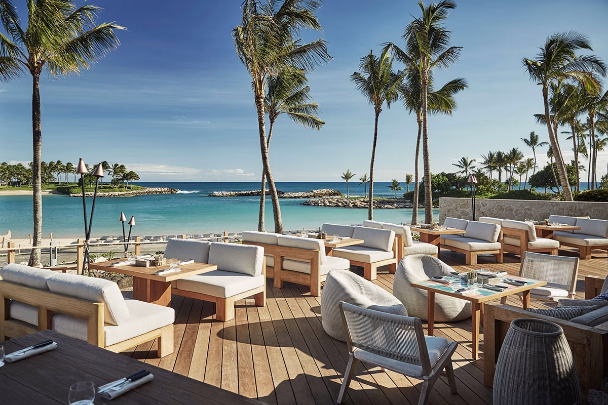 Die weitläufige Holzterrasse des Außenrestaurants im Four Seasons Oahu, ausgestattet mit gemütlichen Loungemöbeln direkt an der sandigen, palmenbesäumten Badebucht