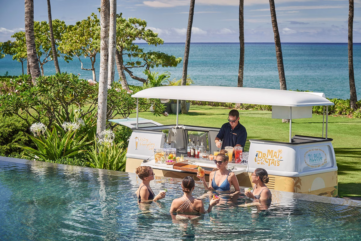 Vier Frauen unterhalten sich im Infinity-Pool des Four Seasons Oahu und bestellen Cocktails an einer umgebauten, stilvollen VW-Bus-Poolbar mit Blick auf den Ozean