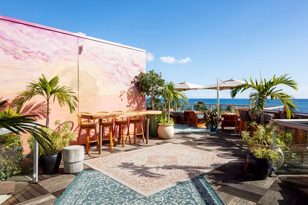 Eine stylische Dachterrasse im Four Seasons Oahu mit Holzboden, gemusterten Teppichen und einer Bar vor einer kunstvoll in Rosatönen bemalten Wand mit Blick auf das weite Meer