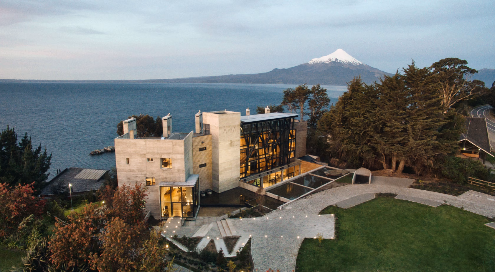 Drohnenaufnahme des Hotel AWA aus Sichtbeton am Ufer eines großen Sees in der Abenddämmerung, im Hintergrund ragt der schneebedeckte, perfekt geformte Vulkan Osorno empor