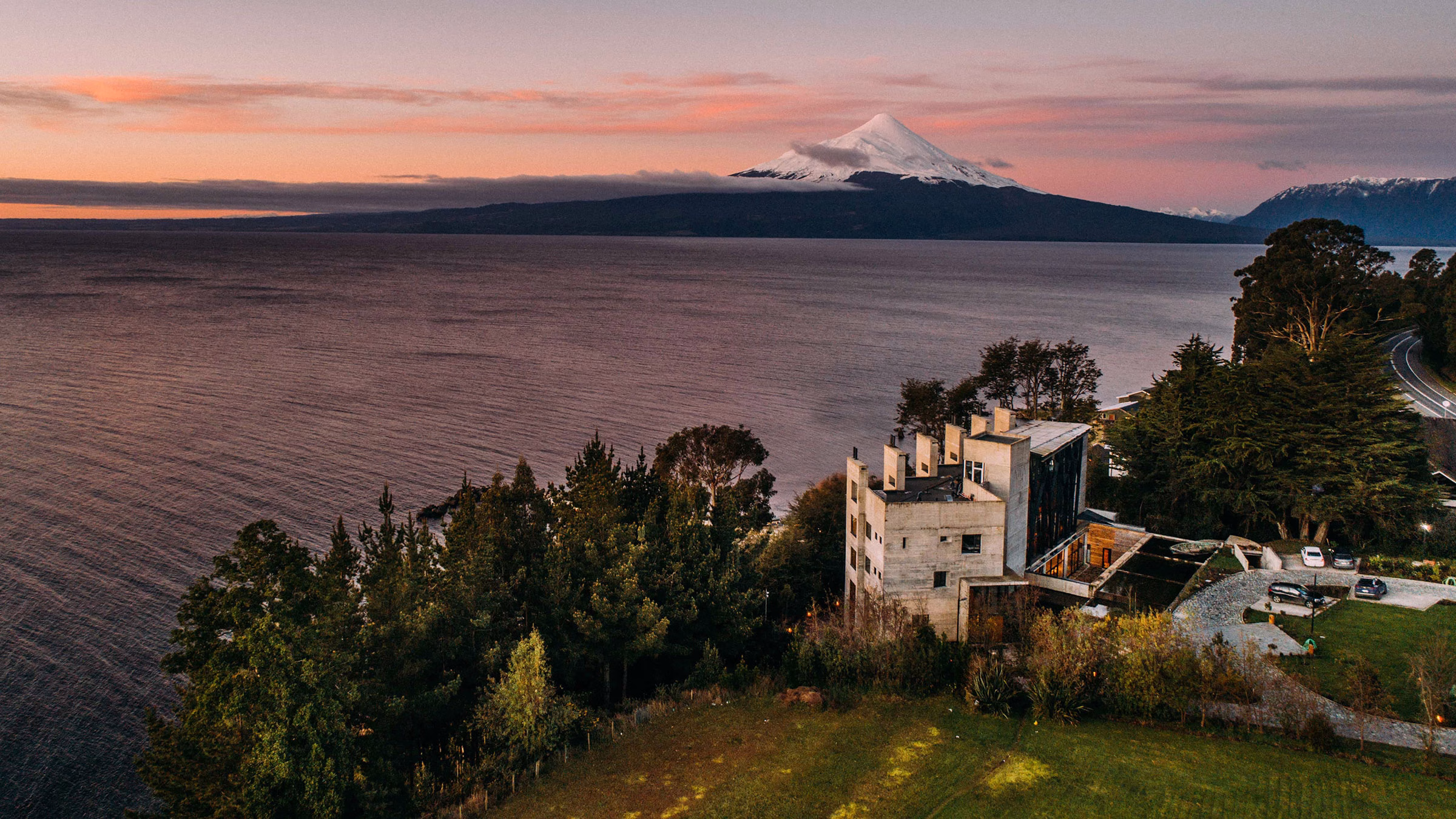 Weite Drohnenperspektive auf das am bewaldeten Seeufer gelegene Hotel AWA bei einem malerischen, rosa-orangefarbenen Sonnenuntergang mit Blick auf den majestätischen Vulkan Osorno