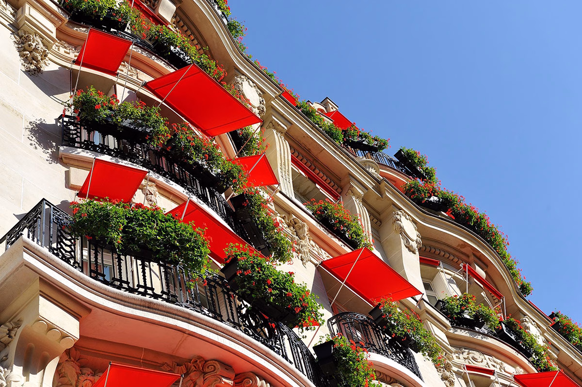 Eine dynamische Nahaufnahme der Pariser Hotelfassade des Plaza Athénée von unten nach oben fotografiert, die die wellenförmigen Balkone mit schwarzen Schmiedeeisengittern, roten Blumenkästen und leuchtend roten Sonnensegeln zeigt