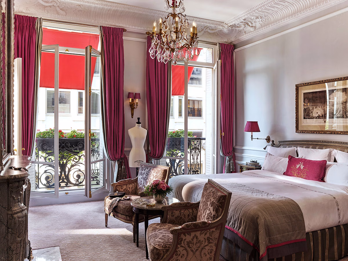Das elegant eingerichtete Schlafzimmer der Haute Couture Suite mit einem großen Doppelbett, einer Schneiderpuppe am Fenster, klassischen Sesseln und edlen, tiefrosa Vorhängen vor den geöffneten Balkontüren
