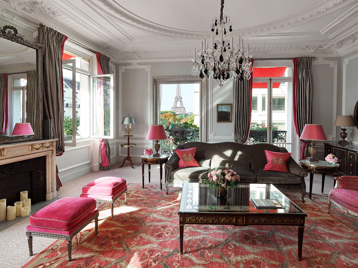 Ein herrschaftlicher Salon in der Haute Couture Suite mit Stuckdecke, einem dunklen Samtsofa mit pinken Kissen, einem verzierten Couchtisch und einem großen Fenster, das den Blick direkt auf den Eiffelturm freigibt