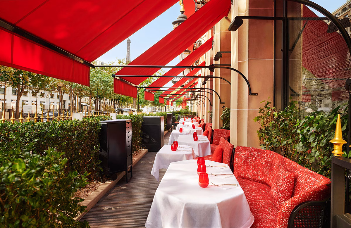 Die Außenterrasse des Restaurants La Terrasse Montaigne an der Straße, geschützt durch eine lange Reihe roter Markisen, ausgestattet mit weißen Tischen und roten Polsterbänken, mit dem Eiffelturm im fernen Hintergrund