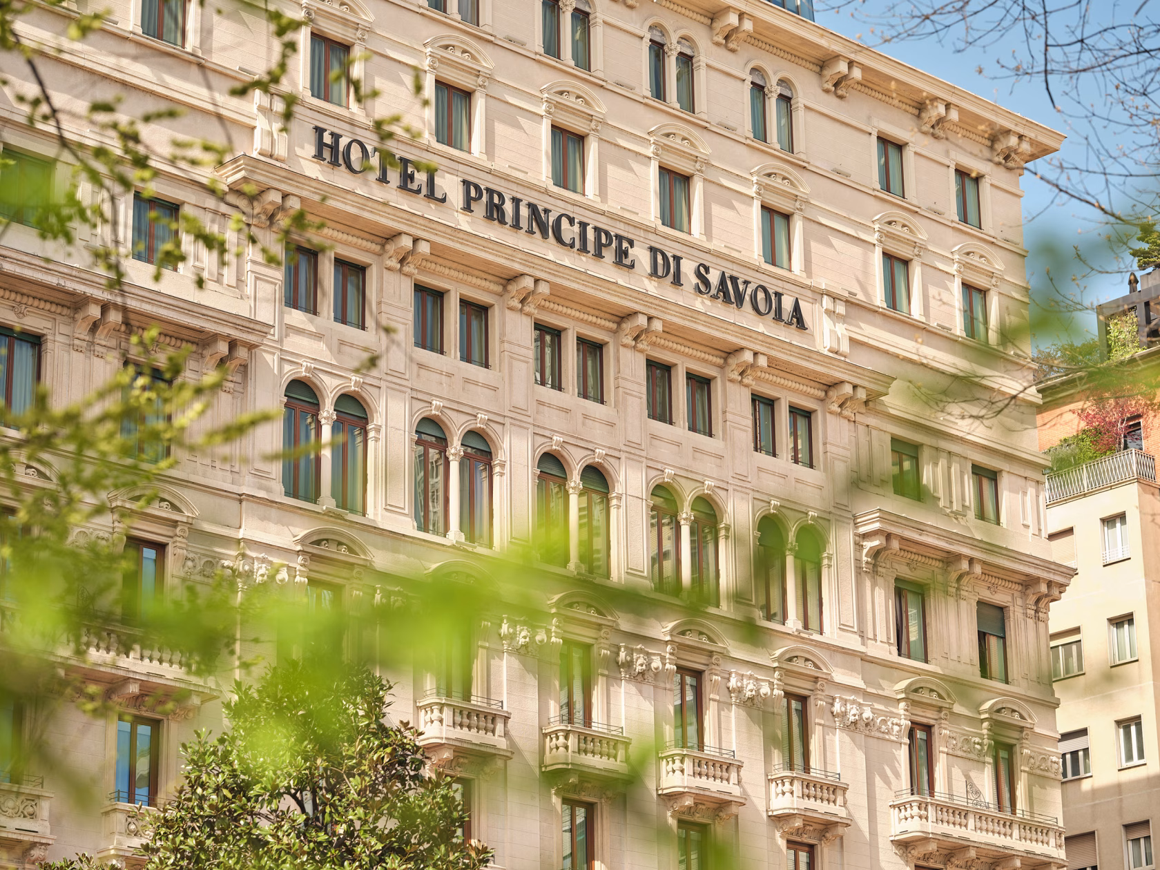 Nahaufnahme der historischen, neoklassizistischen Fassade des Hotel Principe di Savoia in Mailand mit dem großen Schriftzug des Hotelnamens, eingerahmt von frischen, grünen Blättern im Vordergrund
