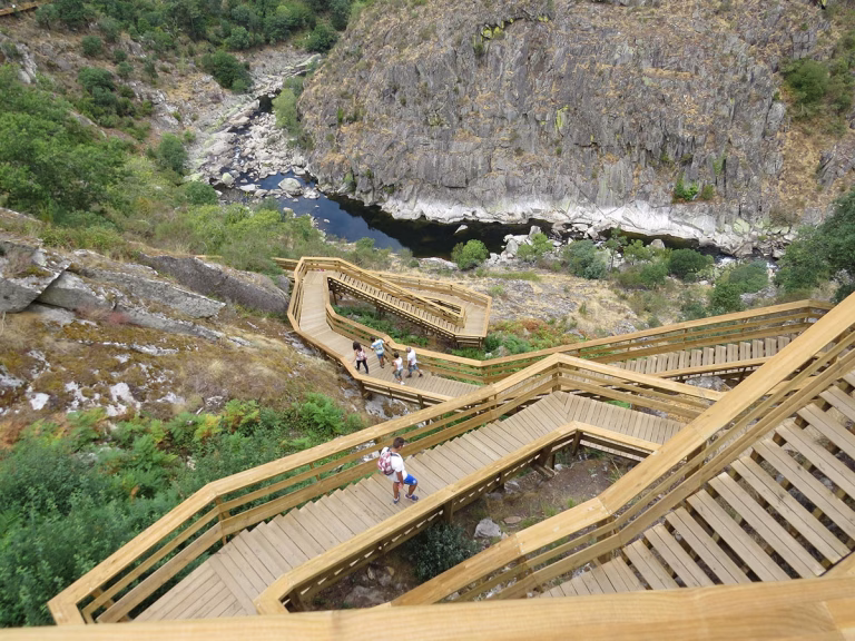 Blick von oben auf die hölzernen Treppenkonstruktionen der Paiva Walkways, die sich im Zickzack an einem steilen Felshang hinabwinden, während Wanderer in Richtung des unten fließenden Paiva-Flusses laufen