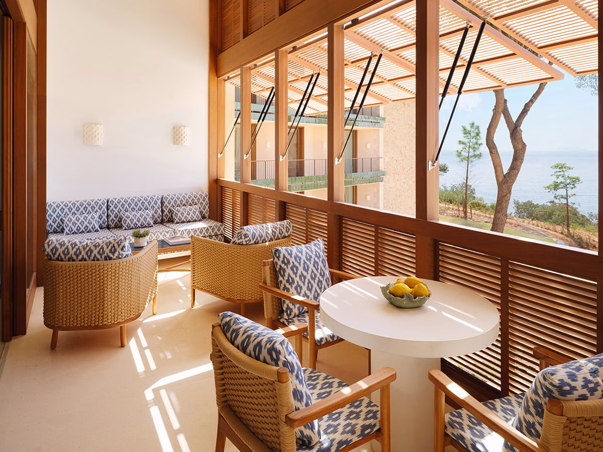 Eine schattige, edle Terrasse im Mandarin Oriental Punta Negra mit blau-weiß gemusterten Loungemöbeln, einem runden Esstisch und hölzernen Lamellenfenstern, die den Blick auf den blauen Ozean freigeben