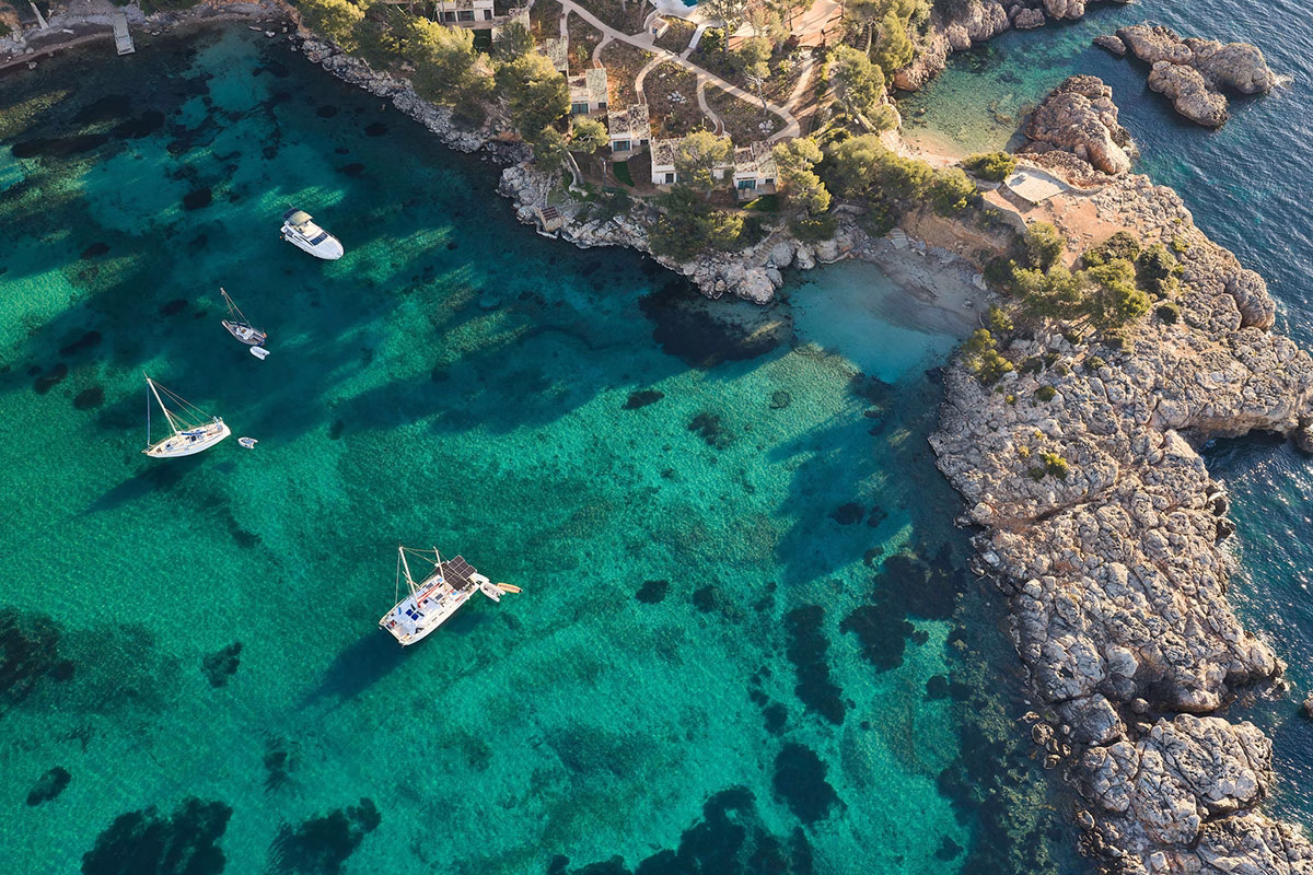 Drohnenaufnahme einer idyllischen, privaten Badebucht am Mandarin Oriental Punta Negra mit glasklarem, smaragdgrünem Wasser, in dem mehrere weiße Yachten und Boote vor einer felsigen Halbinsel ankern