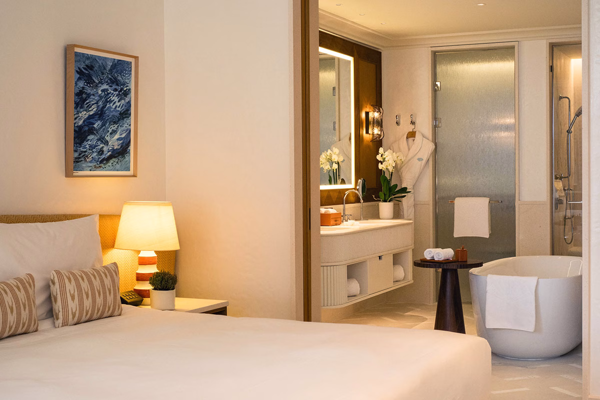 Blick von einem eleganten Hotelbett in das offen gestaltete, helle Badezimmer einer Suite, ausgestattet mit einer freistehenden, ovalen Badewanne, einem beleuchteten Spiegel und edlen Naturstein-Armaturen