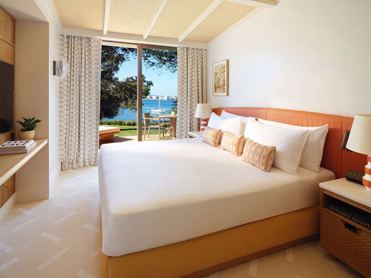 Stilvolles Hotelzimmer mit einem großen Doppelbett, dessen orangefarbenes Kopfteil farbliche Akzente setzt, und einer geöffneten Balkontür, die den Blick auf den blauen Ozean und ein verankertes Boot freigibt