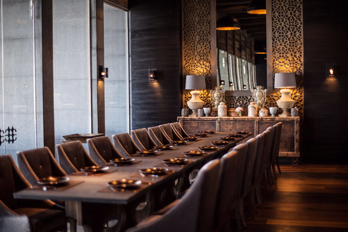 Blick entlang einer langen, minimalistisch eingedeckten Tafel im asiatischen Indochine Restaurant des NIZUC Resort & Spa, akzentuiert durch kunstvolle Holzgitter-Raumteiler und stimmungsvolle Design-Lampen