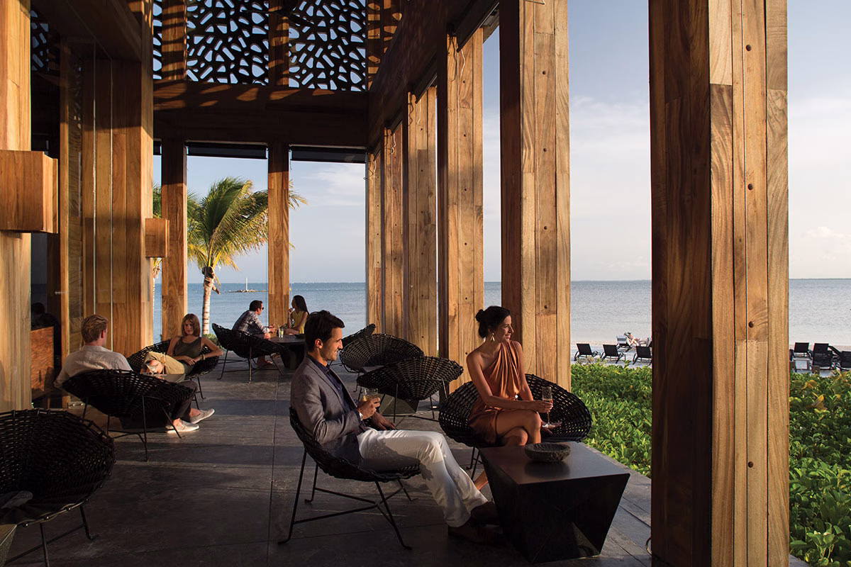Die stilvolle A'Kan Bar im NIZUC Resort & Spa mit einer offenen Architektur aus massiven Holzpfeilern, modernen Flechtsesseln und Hotelgästen, die Drinks bei einem weiten Panoramablick über das Meer genießen