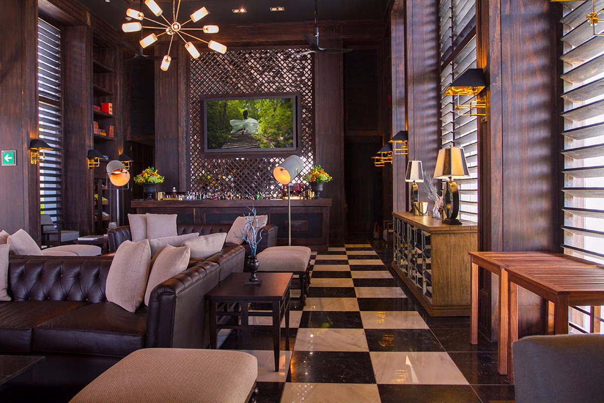 Das edle Interieur der Havana Bar im NIZUC Resort & Spa mit dunkler Holzvertäfelung, Chesterfield-Ledersofas, einem schwarz-weiß karierten Fliesenboden und einer großen Fensterfront mit Holz-Lamellen