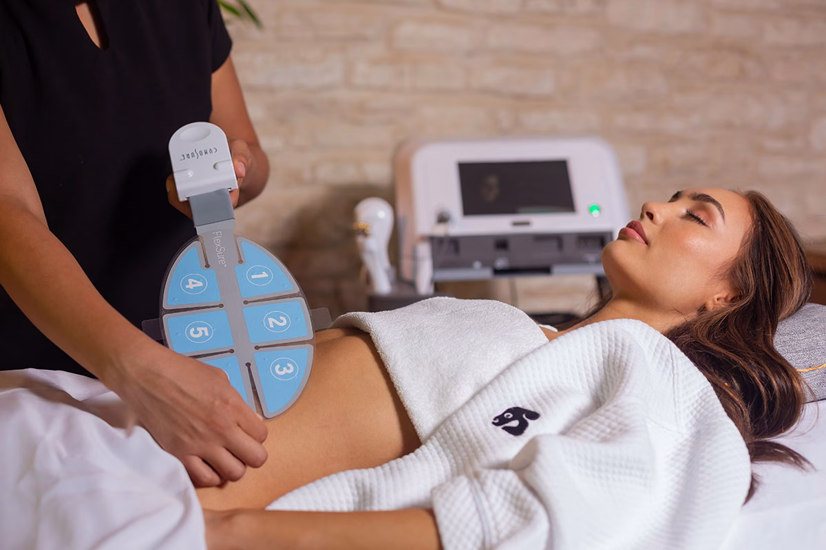 Eine kosmetische Behandlung im NIZUC Med Spa, bei der eine entspannt liegende Frau im weißen Bademantel ein spezielles, blaues Applikations-Pad einer FlexSure-Apparatur auf den Bauch aufgelegt bekommt