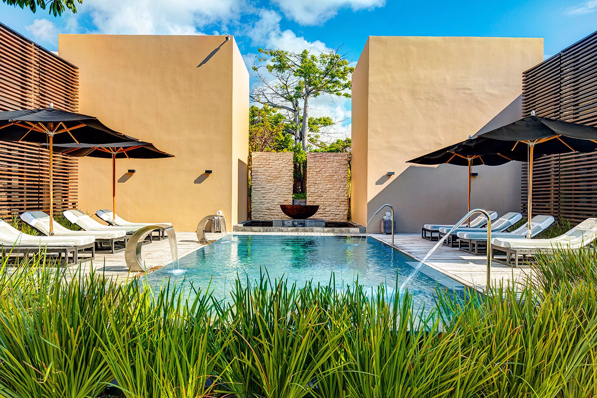 Der exklusive Außenpool des NIZUC Spa by ESPA mit modernen Schwanenhals-Massageduschen, umgeben von hellen Gebäudekuben, schwarzen Sonnenschirmen und gemütlichen Liegen inmitten von dichtem, grünem Ziergras