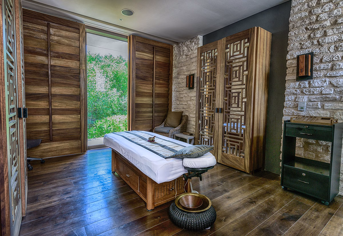 Ein privater Behandlungsraum im NIZUC Spa mit einer edlen Massageliege im Zentrum, umgeben von Wänden aus hellem Naturstein und Schränken aus reich gemasertem Tropenholz sowie einer Schiebetür zum Garten