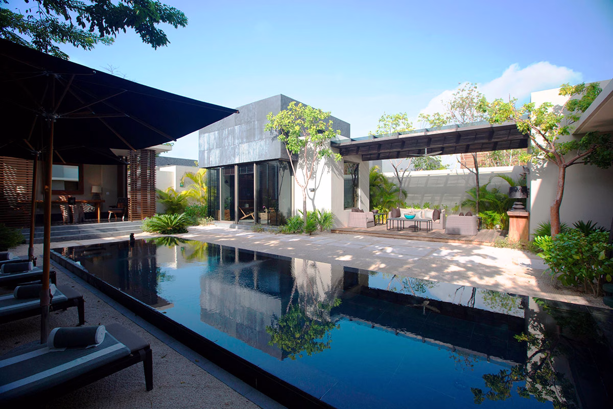 Luxuriöser Außenbereich einer privaten Villa im NIZUC Resort & Spa mit einem großen, spiegelnden Pool, Sonnenliegen unter dunklen Schirmen, einem separaten Loungebereich mit moderner Überdachung und dichter tropischer Bepflanzung
