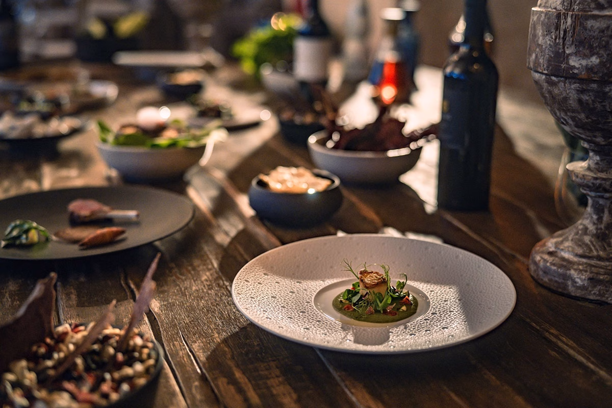 Nahaufnahme eines kunstvoll angerichteten Fine-Dining-Gerichts auf einem weißen Designteller, serviert auf einem rustikalen Holztisch im NIZUC Resort & Spa im warmen, atmosphärischen Abendlicht