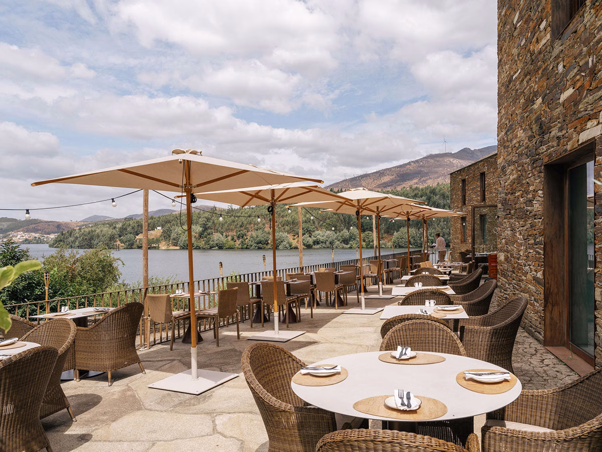 Die weitläufige Außenterrasse des ÀTERRA Restaurants im Octant Douro mit stilvollen Korbstühlen, gedeckten Tischen und hellen Sonnenschirmen direkt am Ufer des Douro-Flusses unter leicht bewölktem Himmel