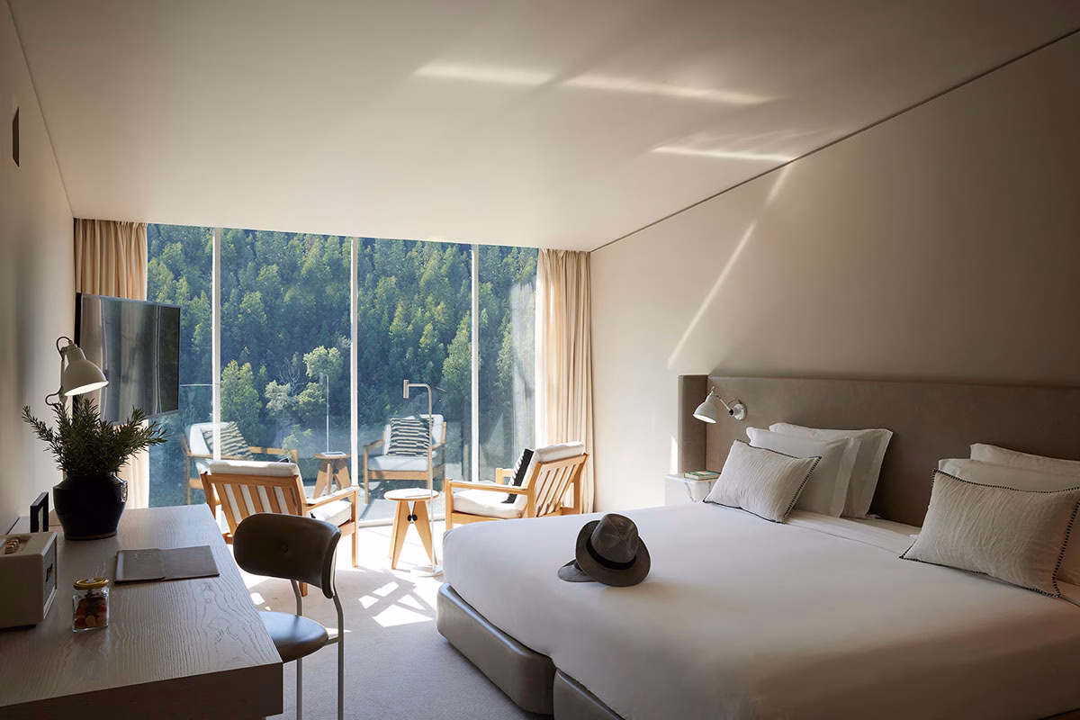 Ein minimalistisch eingerichtetes Deluxe-Zimmer im Octant Douro mit einem großen Doppelbett, hellen Wänden und einer bodentiefen Fensterfront, die den Blick auf den dicht bewaldeten Hang gegenüber freigibt