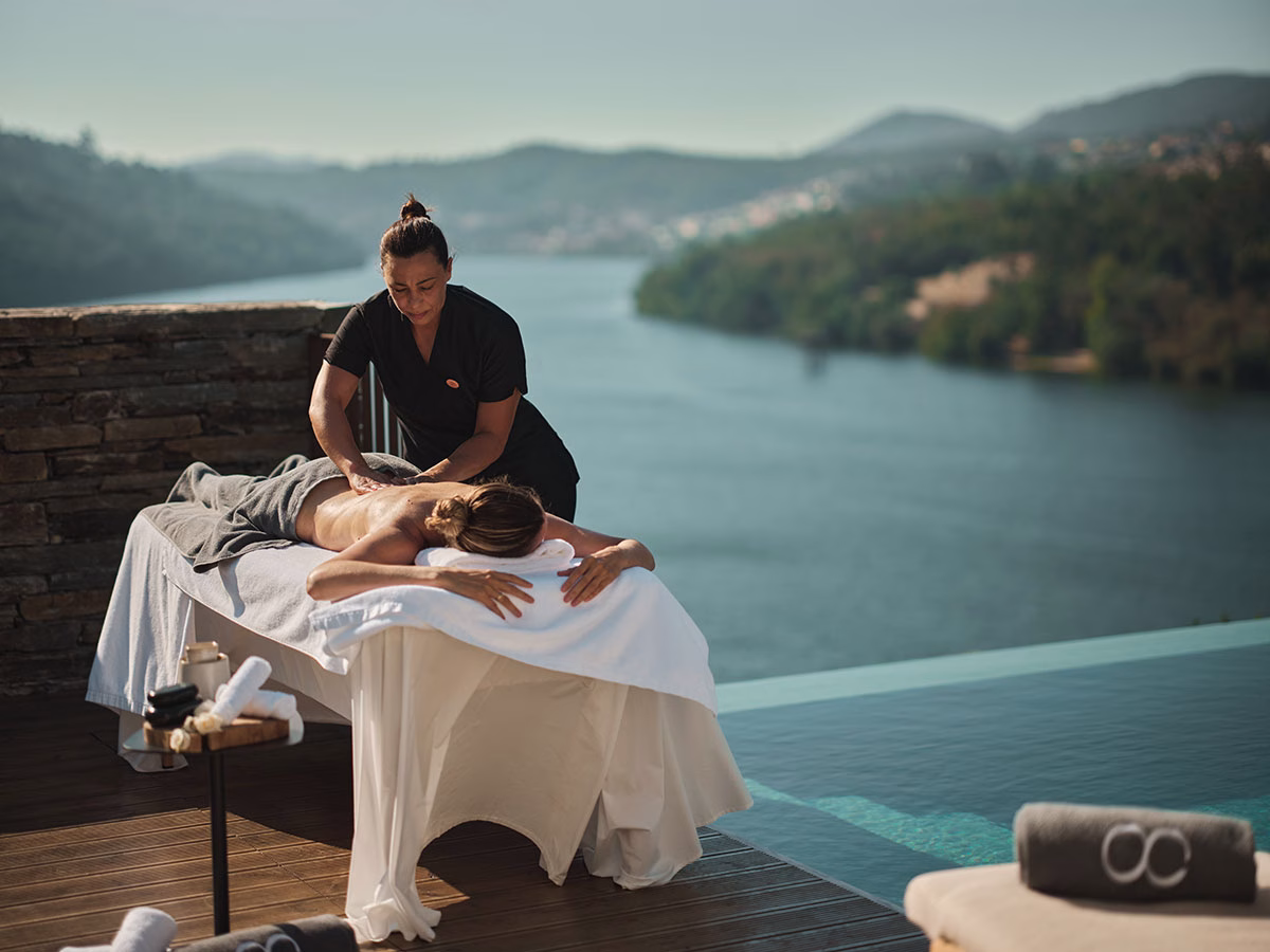 Eine Masseurin bei einer Wellness-Anwendung an einer Kundin auf einer Massageliege im Freien, direkt am Infinity-Pool des Octant Douro mit freiem Blick auf den Douro-Fluss