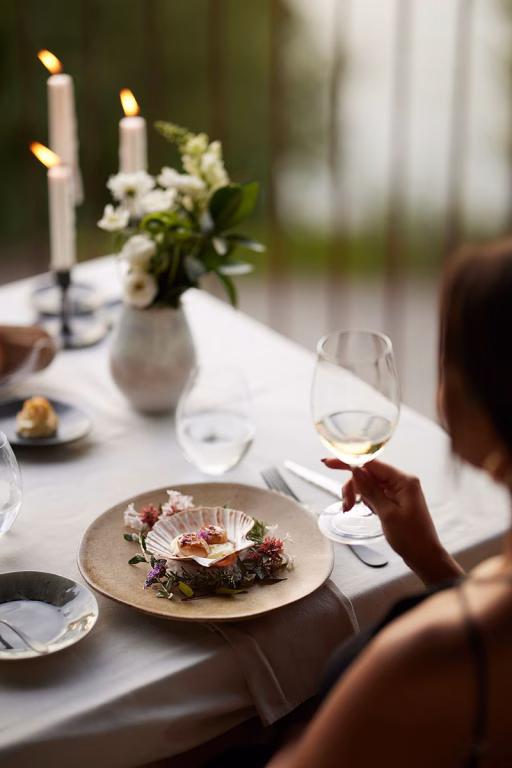 Close-up auf einen elegant gedeckten Tisch im Octant Douro mit Kerzenlicht, feinen Jakobsmuscheln in einer Dekorschale und einer Frau, die ein Glas gekühlten Weißwein hält