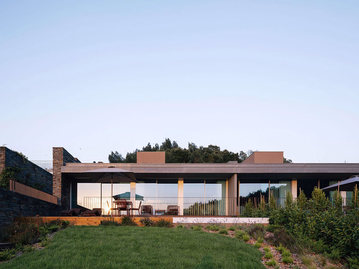 Die moderne Außenfassade der House Collection im Octant Douro im warmen Abendlicht, mit Blick auf eine Rasenfläche, Holzterrassen mit Sonnenschirmen und große Fensterelemente