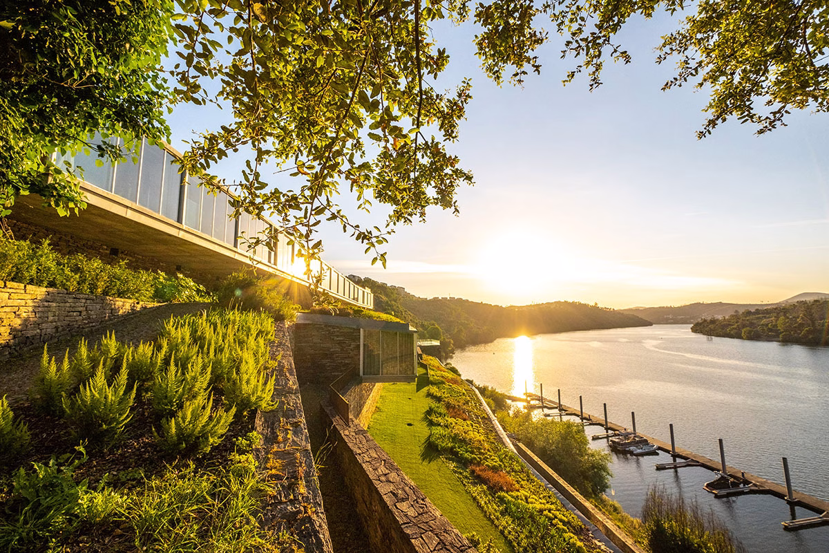 Blick im warmen Abendlicht entlang der terrassierten Steinfassade des Octant Douro, umgeben von grünen Büschen, mit Blick auf den Douro-Fluss und den hoteleigenen Bootsanleger im Hintergrund