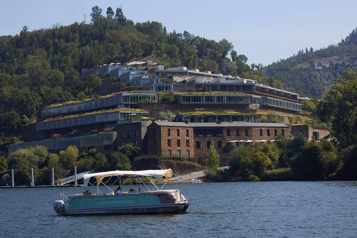 Eine Frontalansicht vom Wasser aus auf das terrassenförmig in den grünen Hang gebaute Octant Douro Hotel aus dunklem Naturstein, während im Vordergrund ein Motorboot auf dem Douro-Fluss fährt