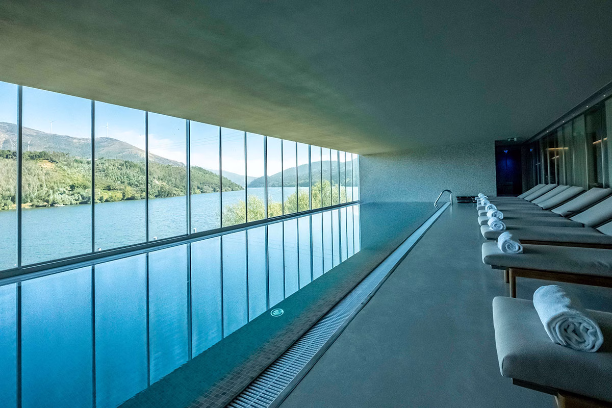 Der weitläufige Innenpool des Octant Douro mit einer lückenlosen Panorama-Glasfront, die den Blick auf den Fluss und die Hügel freigibt, flankiert von einer langen Reihe gepolsterter Holz-Sonnenliegen mit weißen Handtüchern