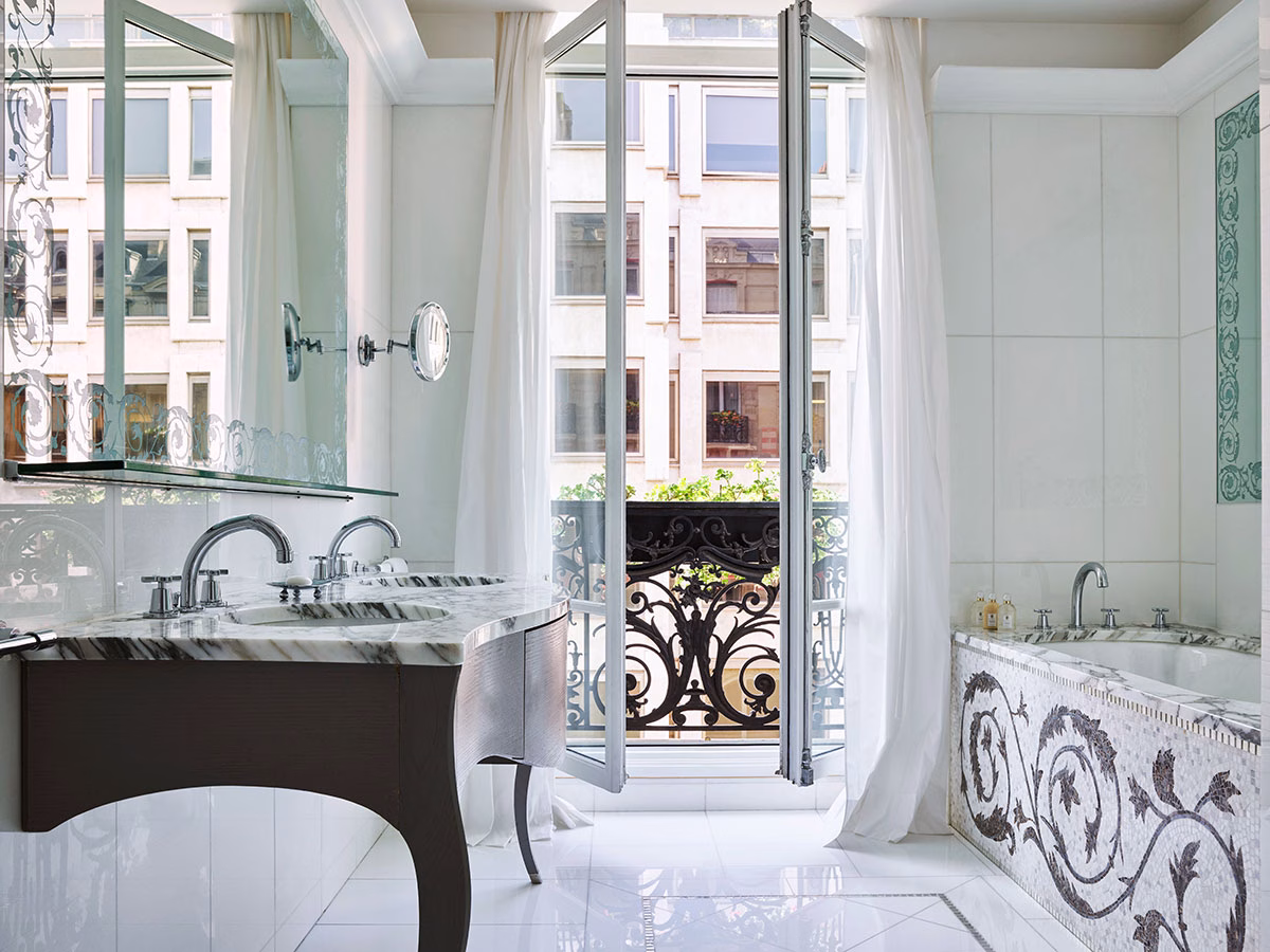 Das lichtdurchflutete, komplett in weißem Marmor gestaltete Badezimmer der Haute Couture Suite 361 mit einer Badewanne inklusive floralem Mosaikdekor, einem eleganten Doppelwaschtisch und geöffneten Flügeltüren zum Pariser Innenhof