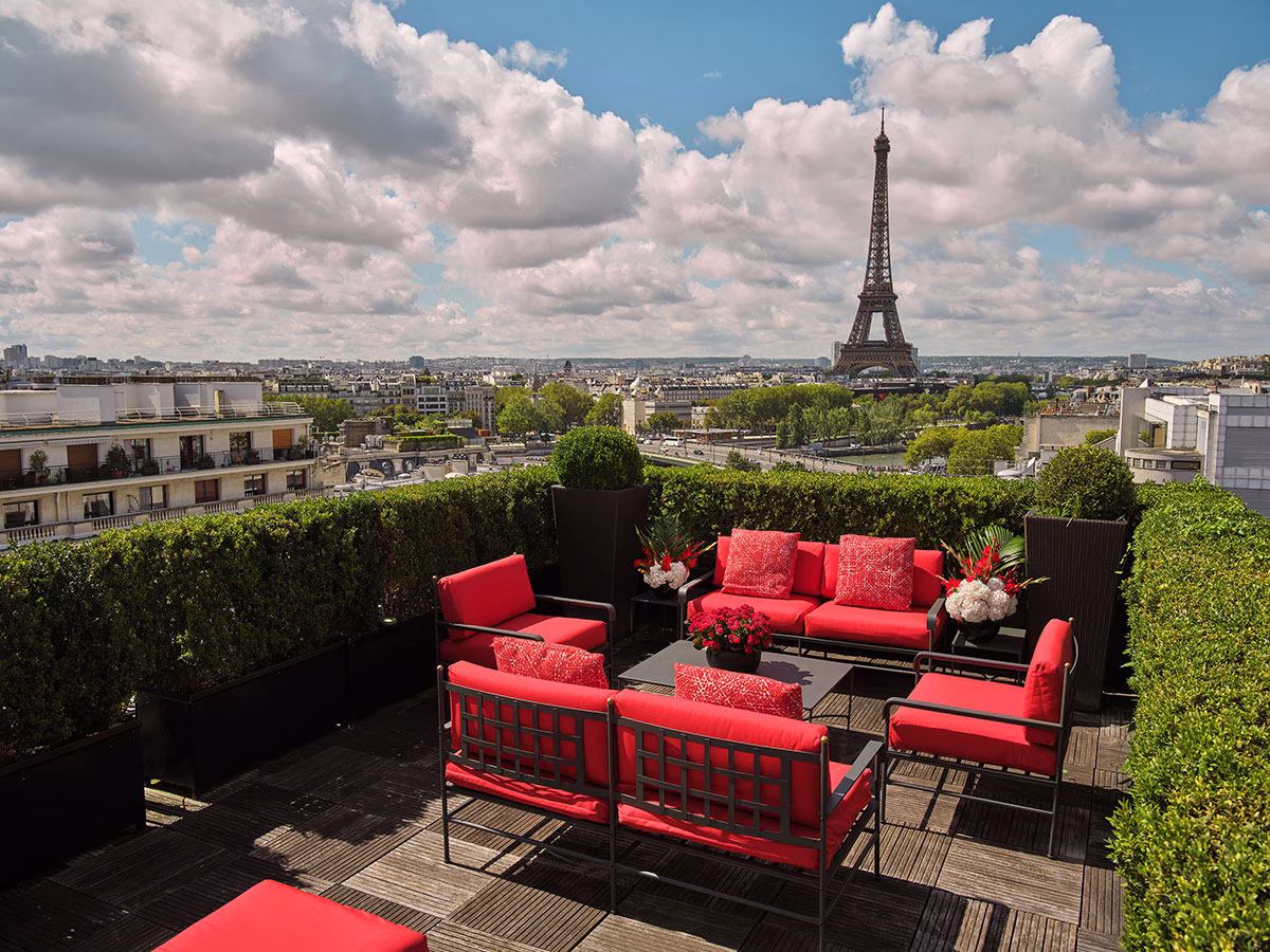 Die exklusive Dachterrasse des Hôtel Plaza Athénée mit gemütlichen Lounge-Möbeln und leuchtend roten Polstern auf Holzdielen, umrahmt von grünen Hecken, mit freiem Blick auf den majestätischen Eiffelturm