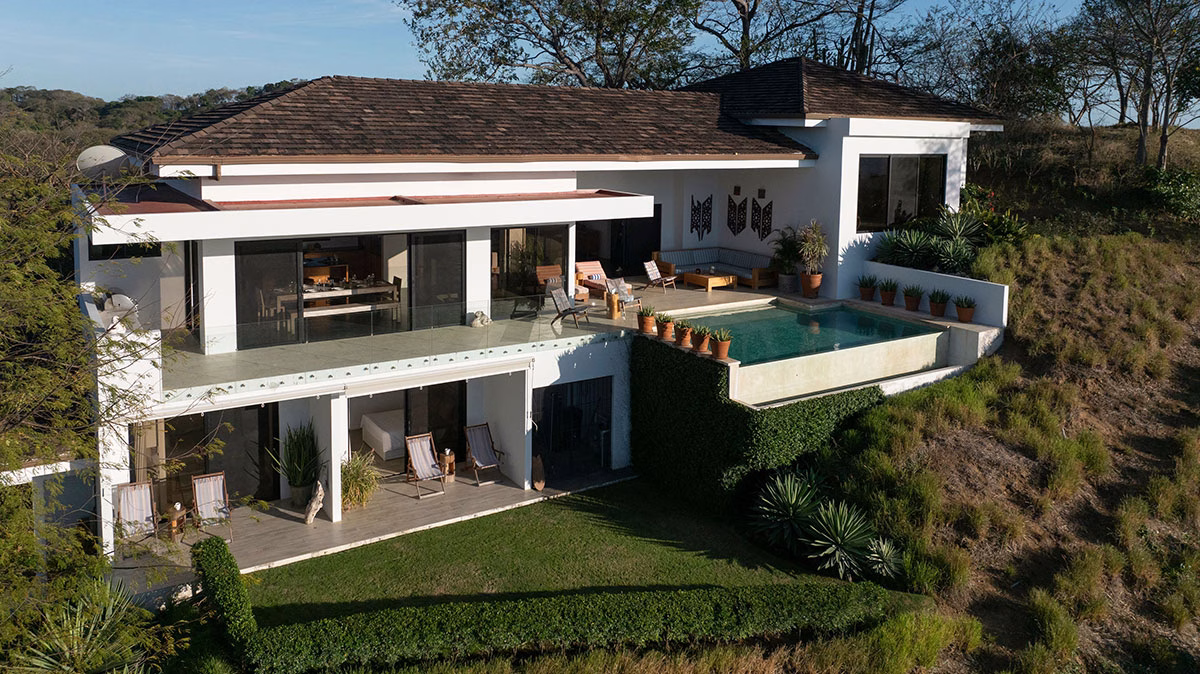 Drohnenperspektive auf eine moderne, weiße Villa der Adventure Collection, die sich an einen grünen Hang schmiegt, komplett mit Infinity-Pool, Balkonterrassen und Meerblick