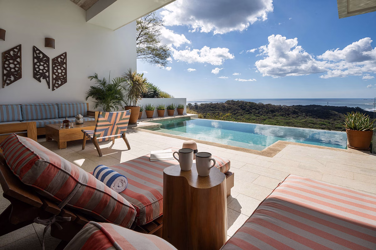 Sonnenterrasse einer Villa im Rancho Santana mit gestreiften Sonnenliegen und zwei Kaffeetassen im Vordergrund, die einen weiten Ausblick über einen Infinity-Pool auf die bewaldeten Hügel und das Meer bietet