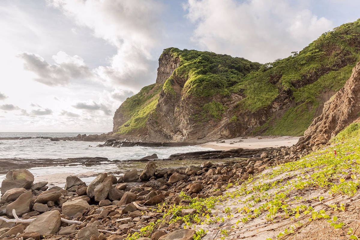 Naturbelassener, felsiger Strandabschnitt der Playa Duna mit einer steilen, grün bewachsenen Klippe, an deren Fuß sich sanfte Wellen brechen