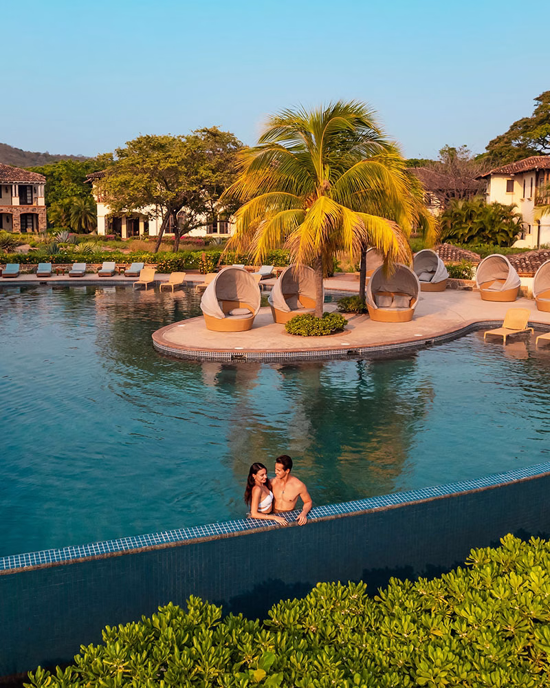 Ein Paar entspannt im großen Infinity-Pool der Residential Suites im Rancho Santana; im Hintergrund befindet sich eine Pool-Insel mit einer Palme und gemütlichen Lounge-Betten