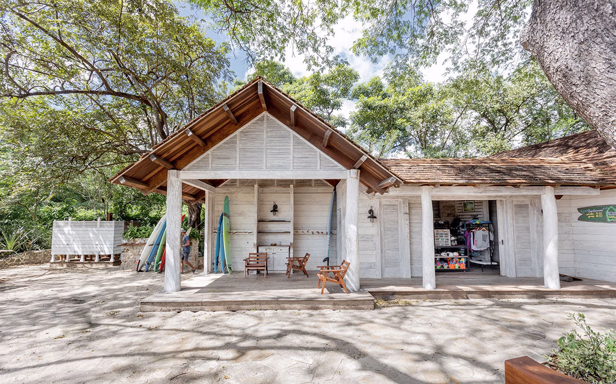 Rustikaler Surf-Shop aus weißem Holz an der Playa Los Perros mit aufgestellten Surfbrettern, schattigen Sitzbänken und dichtem Blätterdach