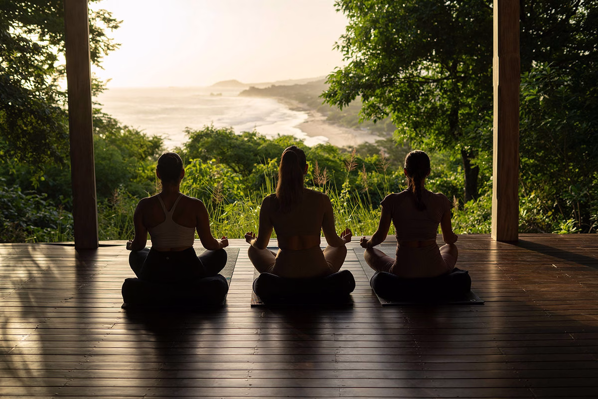 Silhouette von drei Personen beim Meditieren im Yoga-Sitz auf der hölzernen Treetop-Yoga-Palapa mit einem atemberaubenden Panoramablick auf die Küste im Abendlicht