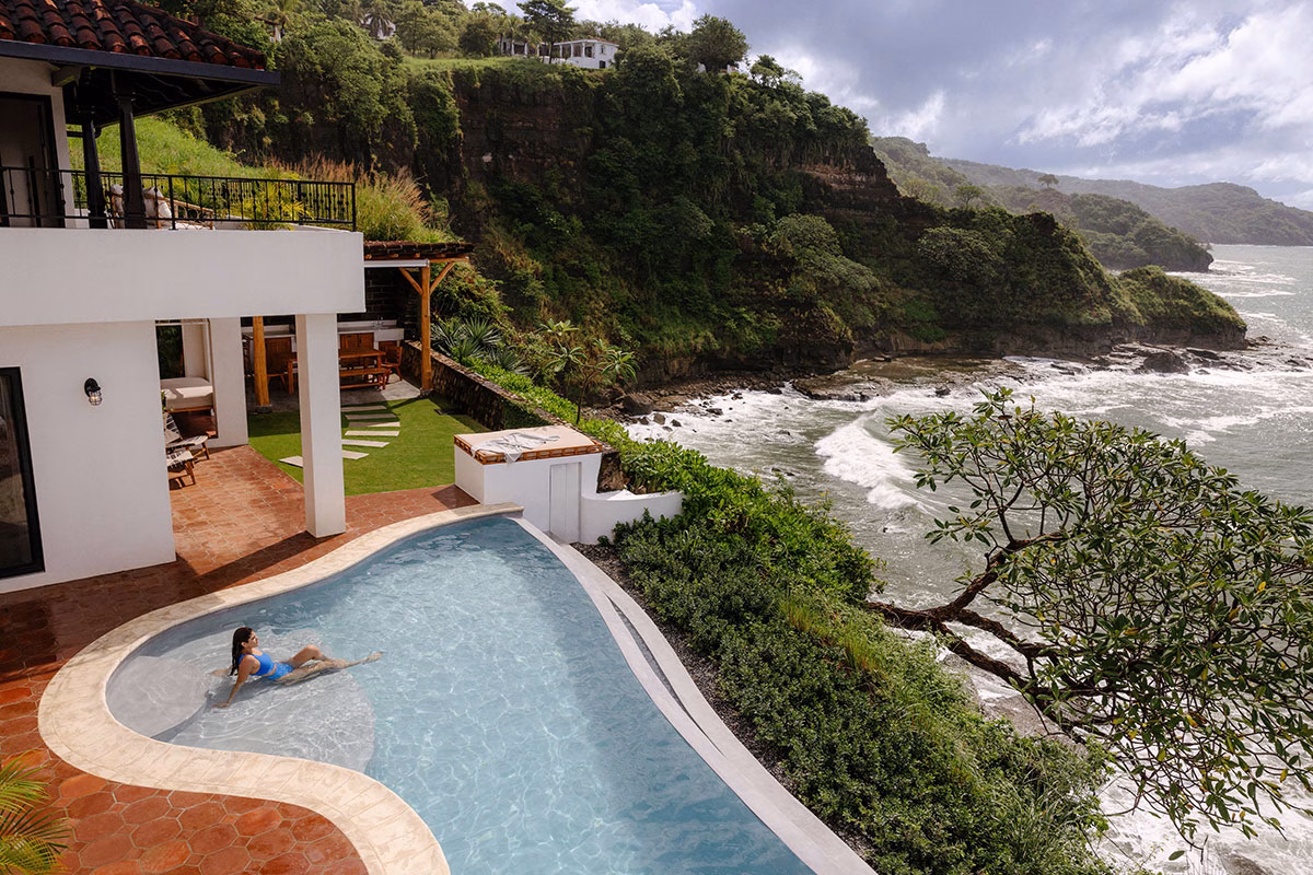 Blick von oben auf die Terrasse einer Klippenvilla, auf der eine Frau im geschwungenen Infinity-Pool entspannt, während unterhalb die Wellen an die felsige Atlantikküste schlagen