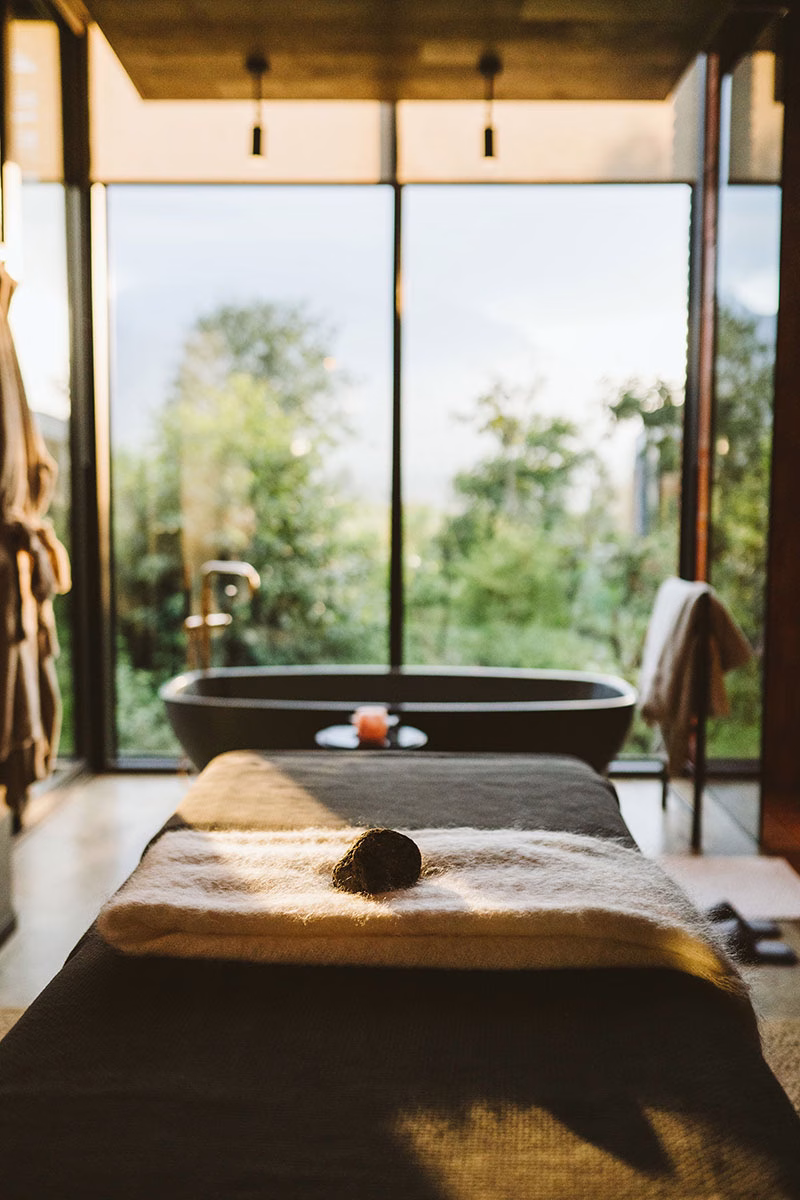 Wellness-Badezimmer in der Singita Kwitonda Lodge mit einer Massageliege im Vordergrund, einer freistehenden Badewanne und einer bodentiefen Glasfront mit Blick in die grüne Natur