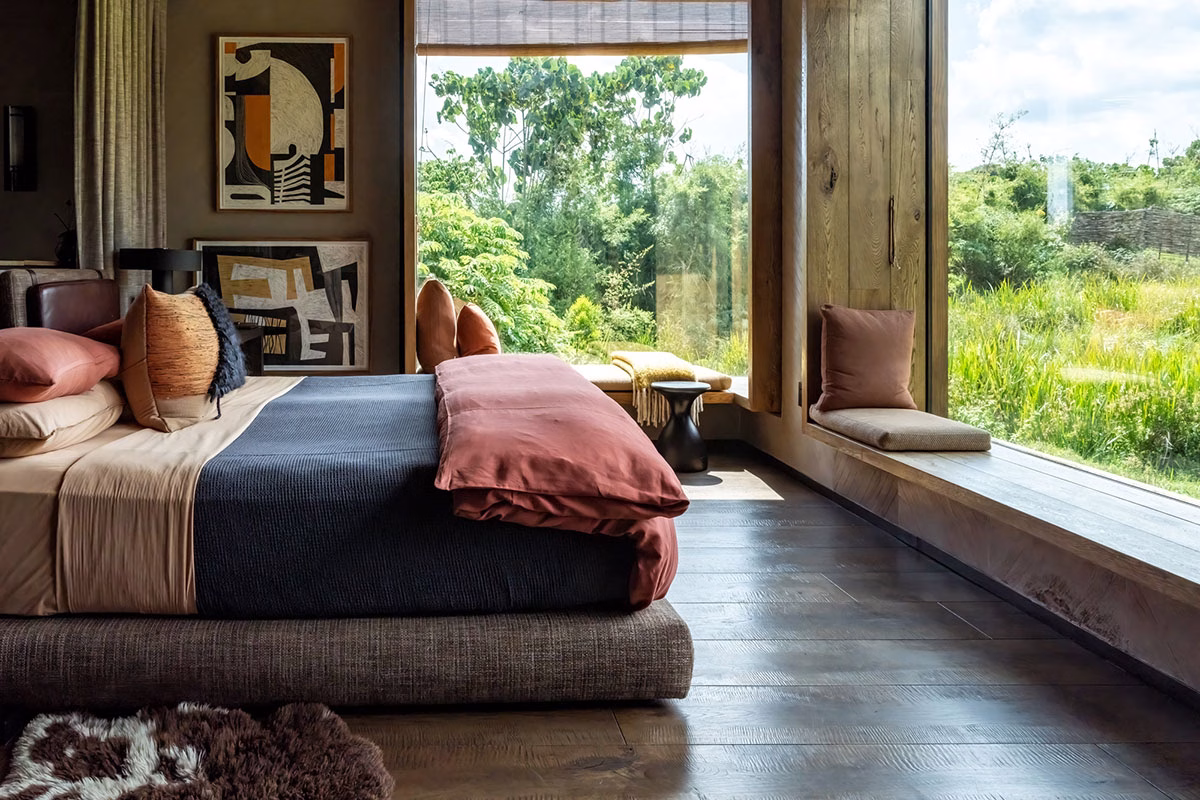 Stilvolles Schlafzimmer in der Singita Kwitonda Lodge mit einem tiefen Bett in Erdtönen, moderner Kunst an der Wand und einer großen, hölzernen Kastenfenster-Sitzbank mit Blick auf das üppige Grün