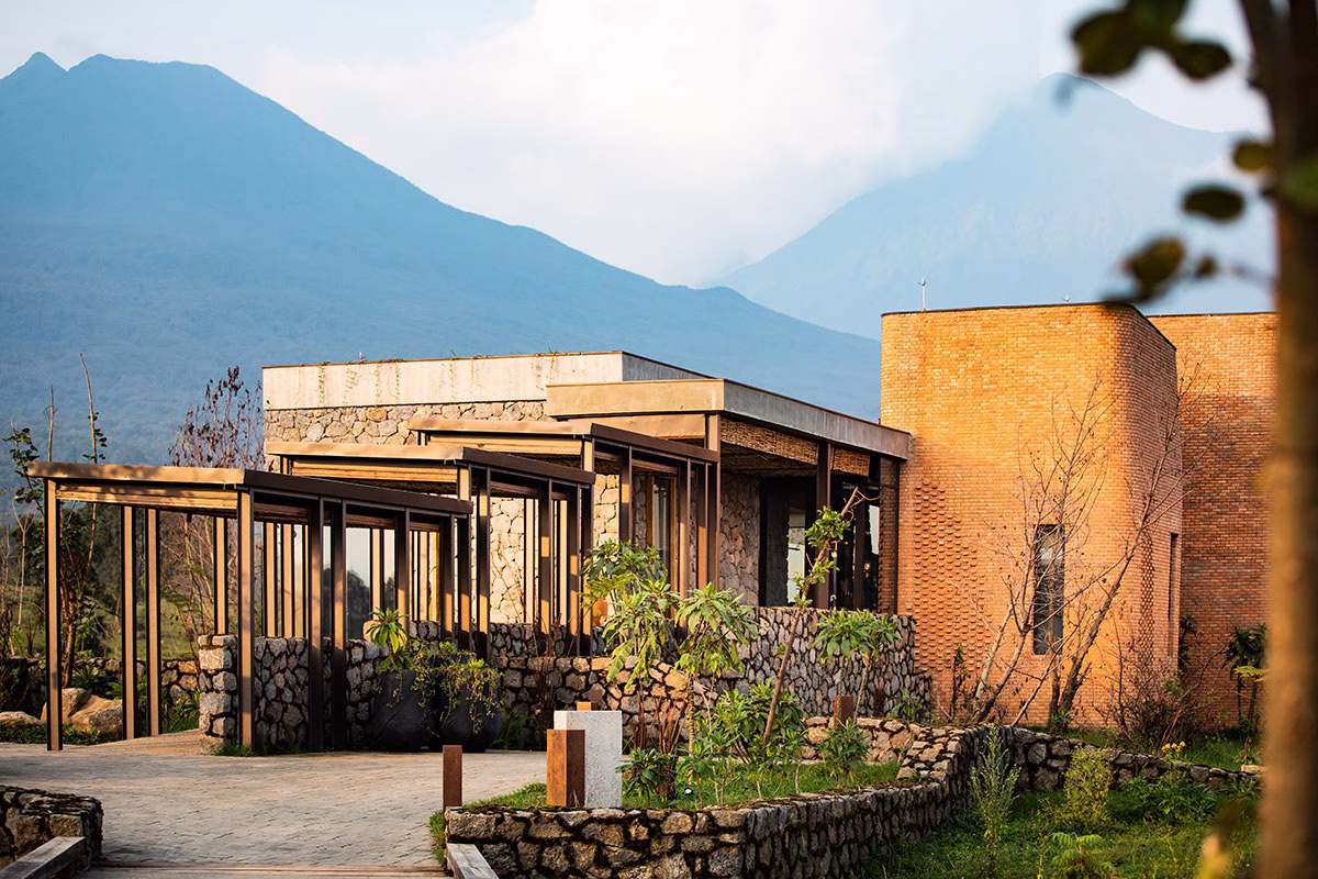 Außenbereich und Architektur der Singita Kwitonda Lodge mit geschwungenen Wegen aus Vulkangestein, rötlichen Backsteinbauten und den nebelverhangenen Virunga-Vulkanen im Hintergrund