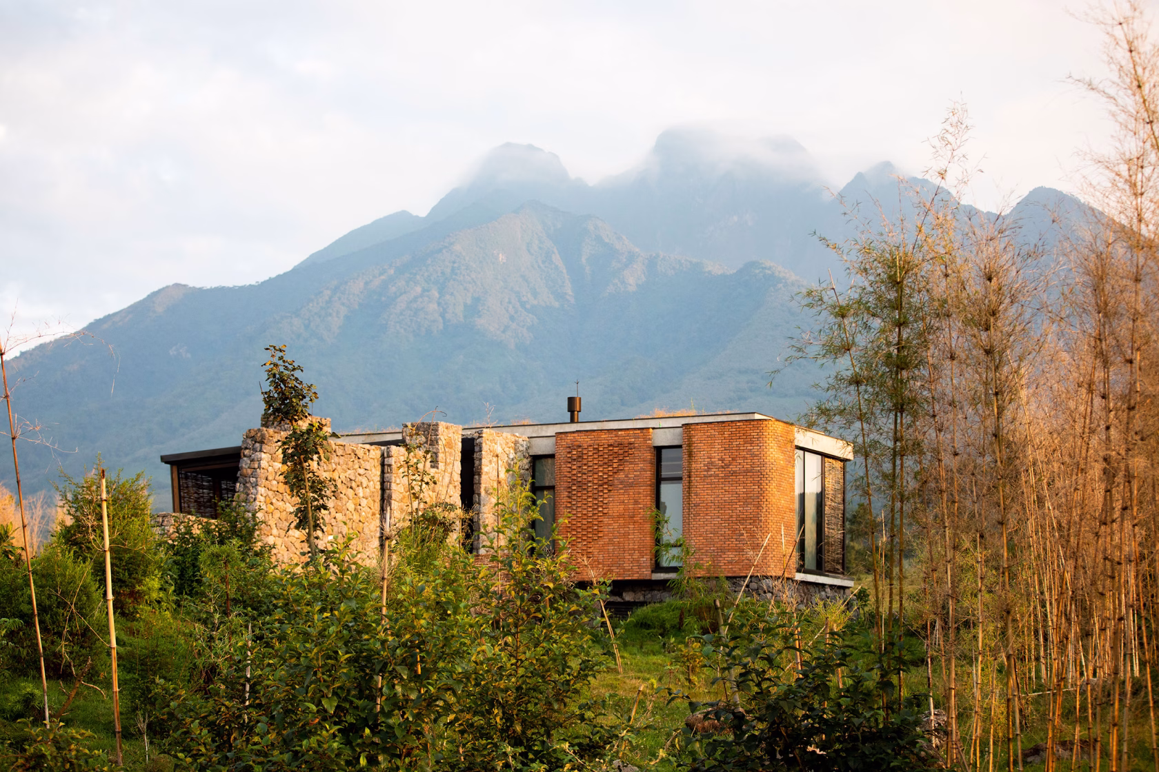 Außenansicht einer exklusiven Ein-Zimmer-Suite der Singita Kwitonda Lodge aus Backstein und Naturstein, umgeben von dichtem afrikanischen Buschland unter wolkenverhangenen Bergen