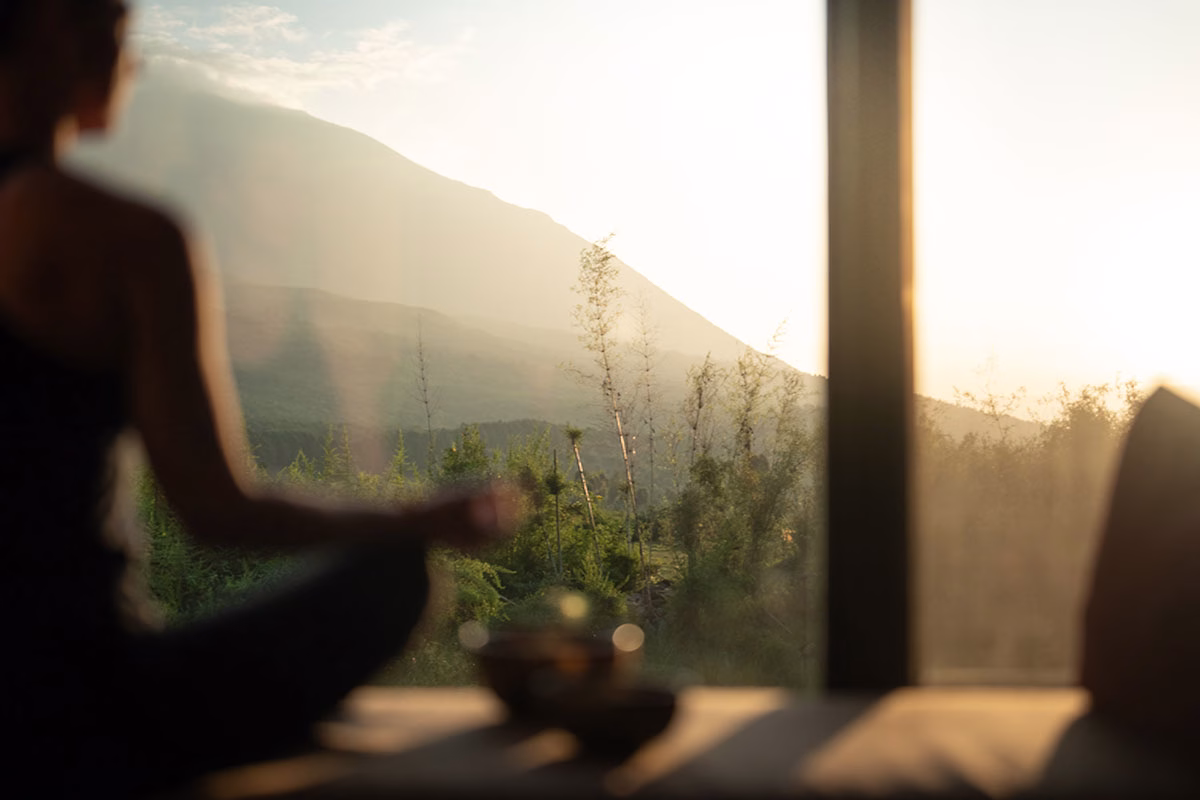 Silhouette einer Person im Meditationssitz (Lotussitz) im sanften Gegenlicht der Morgensonne, die durch eine große Fensterfront blickt, im Hintergrund die nebelverhangenen Hänge des Volcanoes-Nationalparks