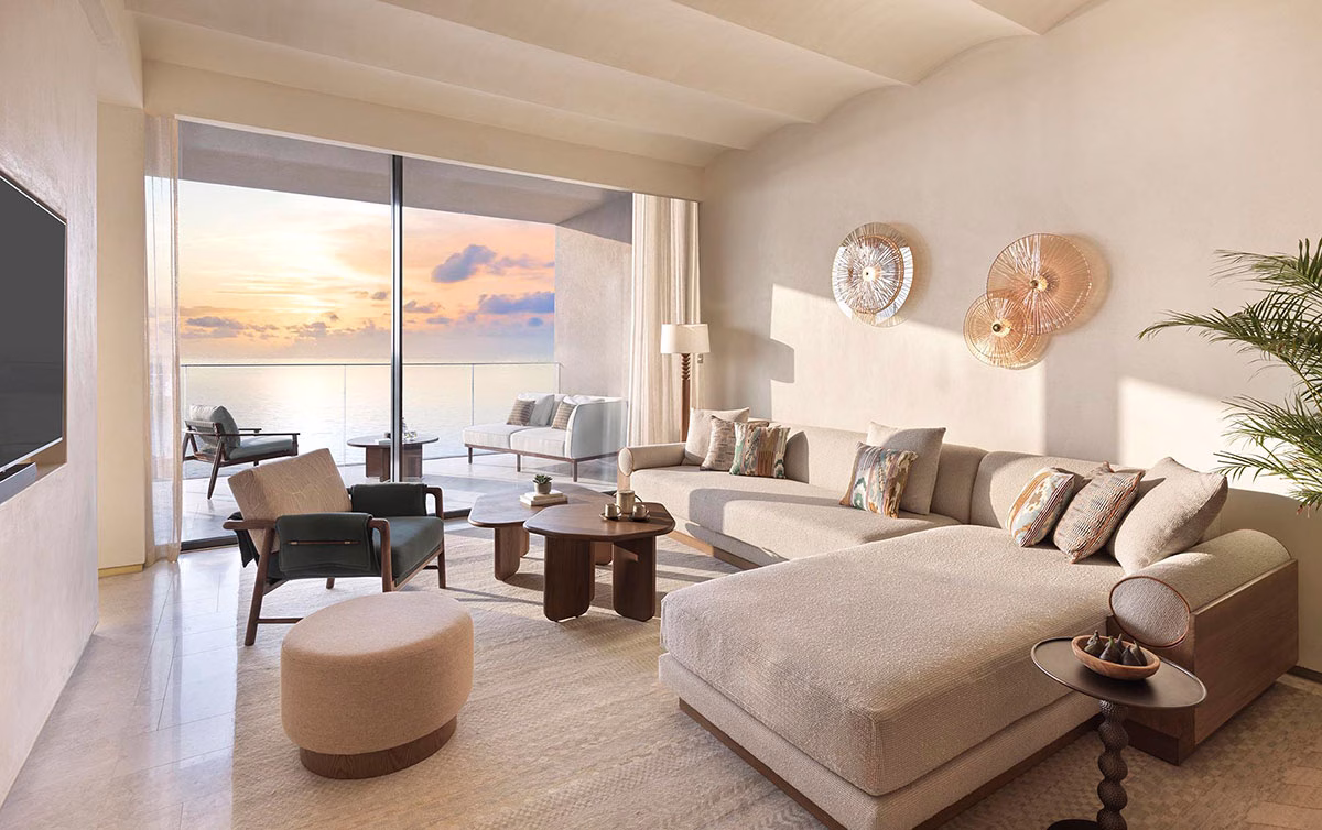 Stilvoller Wohnbereich der Breeze Suite mit großem Sofa, modernen Holztischen und einem Panoramablick auf das Meer durch die verglaste Front, die auf einen Balkon führt