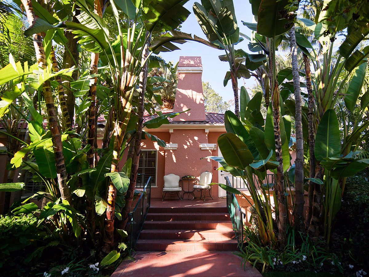 Eingangsbereich von Bungalow 4A im The Beverly Hills Hotel mit einer ziegelroten Treppe, die zu einer Terrasse mit zwei eleganten Sesseln führt, umgeben von dicht gewachsenen Bananenstauden vor der rosafarbenen Fassade