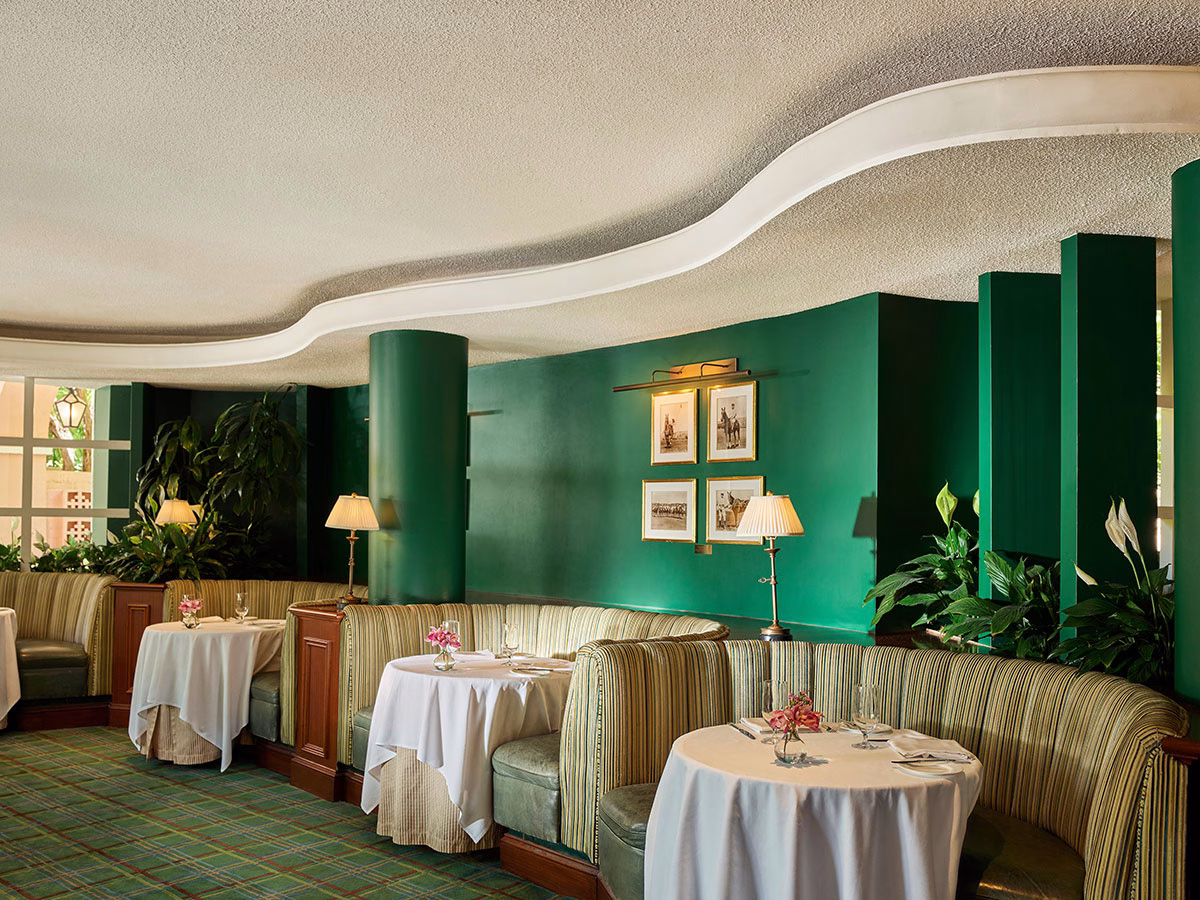 Der elegante Innenbereich der Polo Lounge mit geschwungenen, tiefgrünen Wänden, klassischen Sitzecken mit gestreiften Polstern, weiß eingedeckten Tischen und einem grün-karierten Teppichboden unter einer wellenförmigen Decke