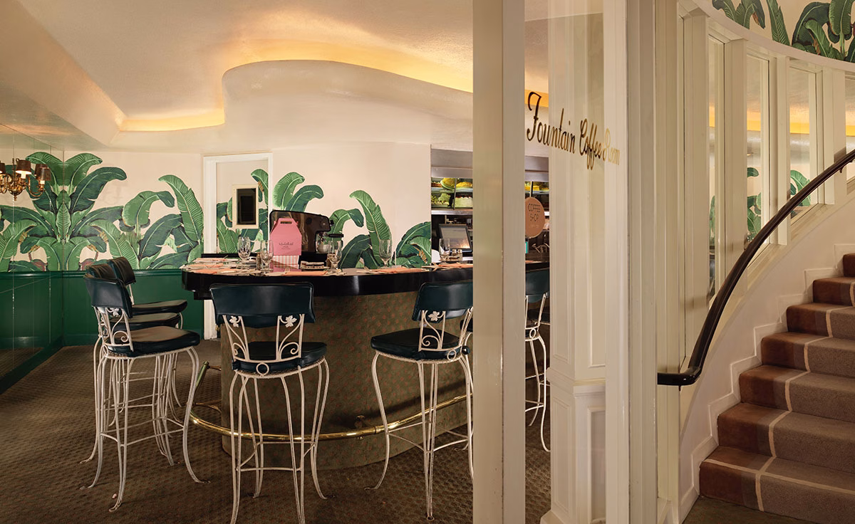 Blick in den historischen The Fountain Coffee Room mit einer geschwungenen, dunklen Theke und weißen Barhockern mit dunkelgrünen Polstern vor einer Wand mit der ikonischen Bananenblatt-Tapete neben einer Treppe